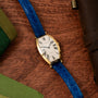 The Cartier Tonneau XL gold watch with a blue leather band rests on a wooden surface, surrounded by green and brown items.