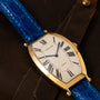 The Cartier Tonneau XL watch features Roman numerals and a blue leather strap, displayed on a brown leather surface.
