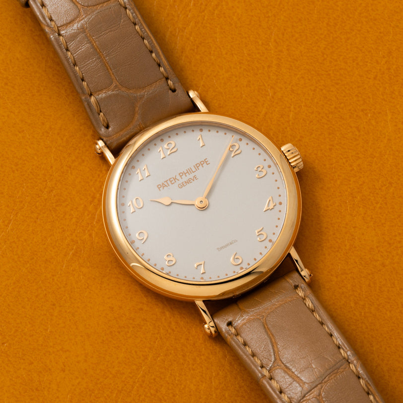A Patek Philippe Calatrava gold wristwatch with a brown leather strap displayed on a mustard yellow background.
