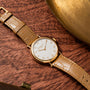 The Patek Philippe Calatrava, featuring a gold case and tan leather strap, rests on a wooden surface beside a brass object.