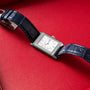 The Jaeger-LeCoultre Reverso Classique Ladies, a rectangular silver watch by Jaeger-LeCoultre with a blue leather strap, displayed on a red leather surface.