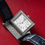 The Jaeger-LeCoultre Reverso Classique Ladies, a rectangular silver watch with a blue strap, diamond bezel, and open caseback, is displayed on a red surface.