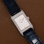 The Jaeger-LeCoultre Reverso Classique Ladies watch features a rectangular silver case, blue leather strap, and diamond accents, elegantly displayed on a brown leather surface.