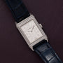 The Jaeger-LeCoultre Reverso Classique Ladies, a rectangular silver watch with diamond accents and blue leather strap, is displayed on a dark purple textured surface.
