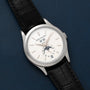 A Patek Philippe Annual Calendar Moonphase watch in silver with a black leather strap, featuring date and moonphase display, placed on a dark blue surface.