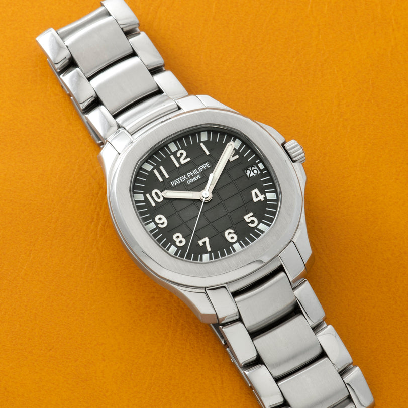 Patek Philippe Aquanaut silver wristwatch with a black dial and metal band, displayed on an orange surface.