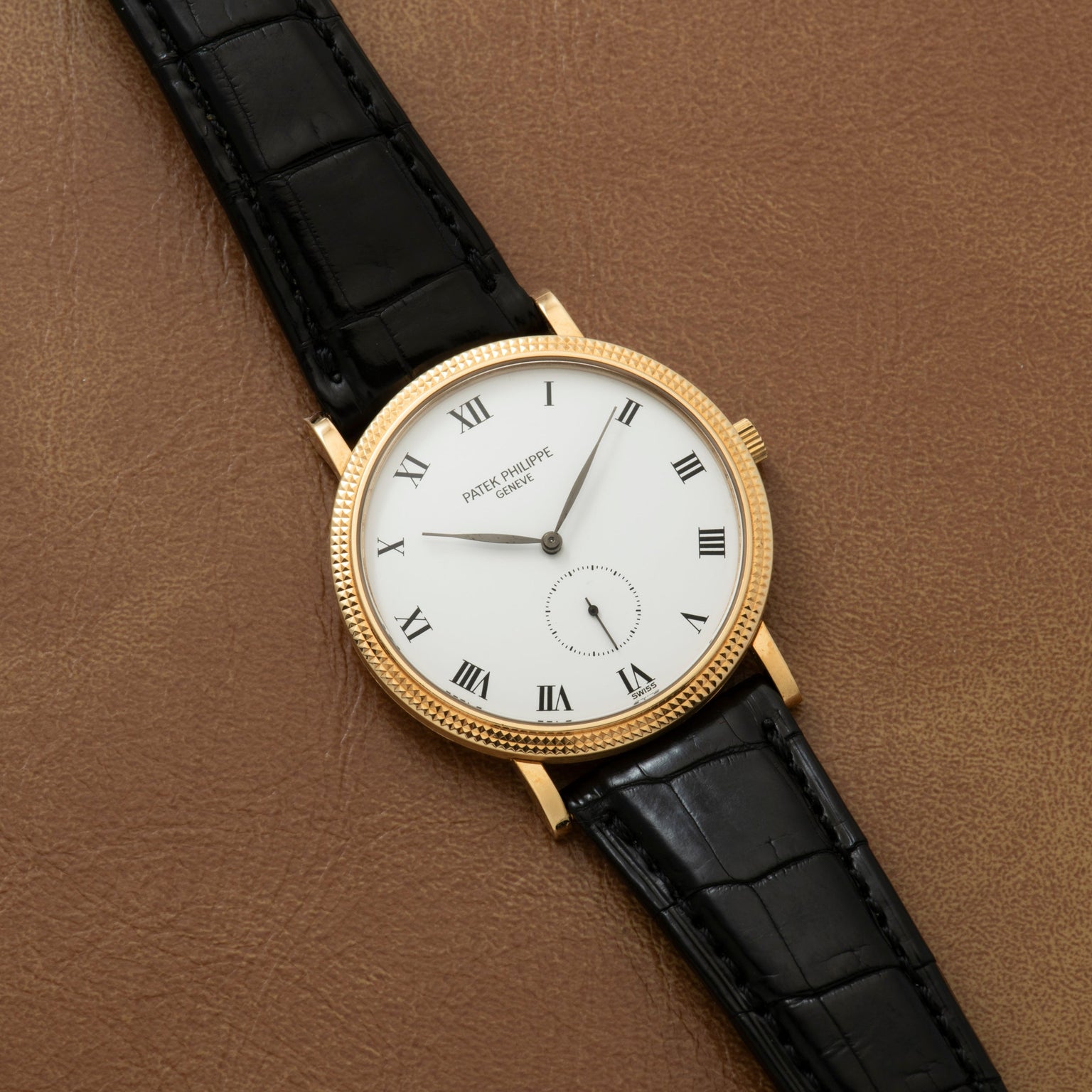 The Patek Philippe Calatrava features a white dial with Roman numerals and a black leather strap, displayed elegantly on a brown surface.