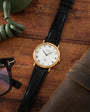A Patek Philippe Calatrava gold watch with a black leather strap rests on a wooden surface beside glasses and a coiled green cord.