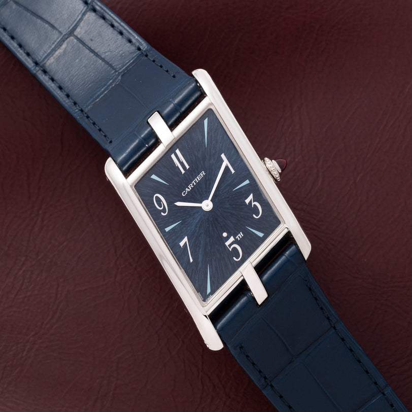 The Cartier Privé Collection Tank Asymétrique New York Edition, a rectangular watch with a blue strap and blue dial, rests on a burgundy leather surface.