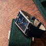 A Cartier Privé Collection Tank Asymétrique New York Edition wristwatch with a square blue face and leather strap rests on a green case atop a wooden surface.
