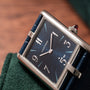 The Cartier Privé Collection Tank Asymétrique New York Edition, featuring a rectangular design with a blue dial and Roman numerals, rests on a green textured surface.