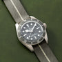 A TUDOR Black Bay 58 Silver dive watch with a black dial and fabric strap is displayed on a textured green surface.