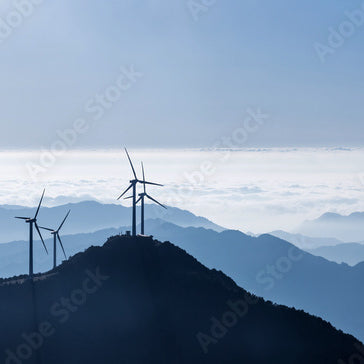 Wind turbines on a mountain ridge with misty hills and clouds in the background under a clear sky.