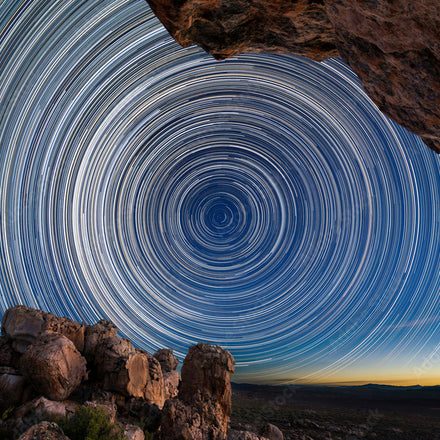 Star trails form circular patterns in the night sky above rocky terrain and a distant landscape.