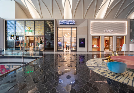 Mall with Rolex, Watches of Switzerland, and Cartier storefronts, glossy floor, and colorful seating in foreground.