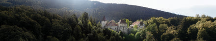 A castle with red rooftops nestled among dense green trees and hills under a cloudy sky.