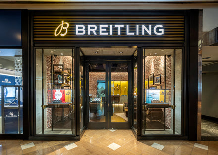 Breitling store entrance with illuminated logo, glass doors, and watch displays inside.