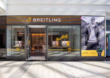 Front view of a Breitling store with glass doors, yellow logo, and watch advertisements displayed.