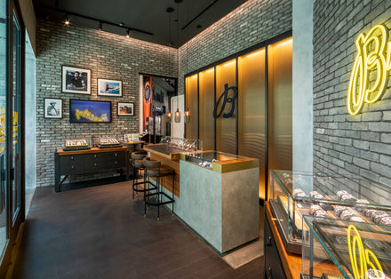 Modern watch store with glass display cases, brick walls, framed photos, and a neon yellow sign on the right.