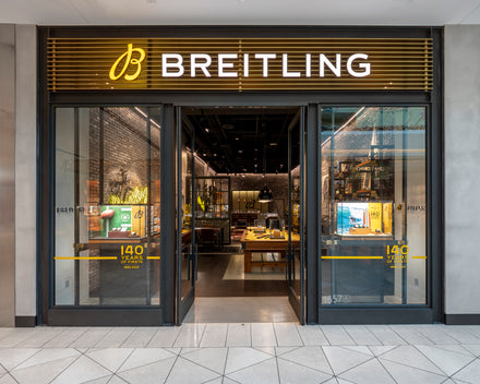 Breitling store entrance with glass doors, yellow signage, and watches displayed inside.