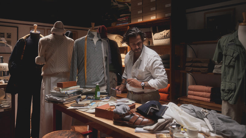 Davide Baroncini stands in a crowded clothing workroom behind a bench covered with items of clothing, accessories, and papers