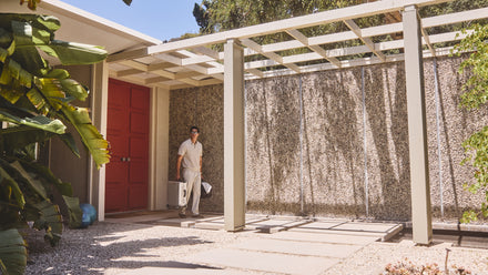 A person carrying shopping bags walks outside a mid-century modern house with red double doors.