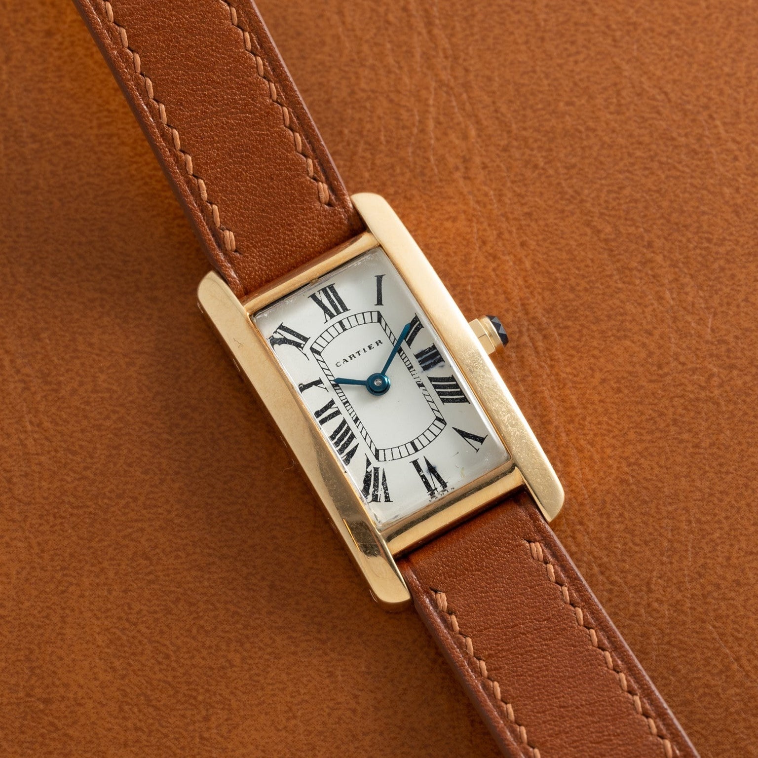 Cartier Tank Cintrée Ladies watch: rectangular gold case, brown leather strap on brown leather surface, Roman numerals on white dial.
