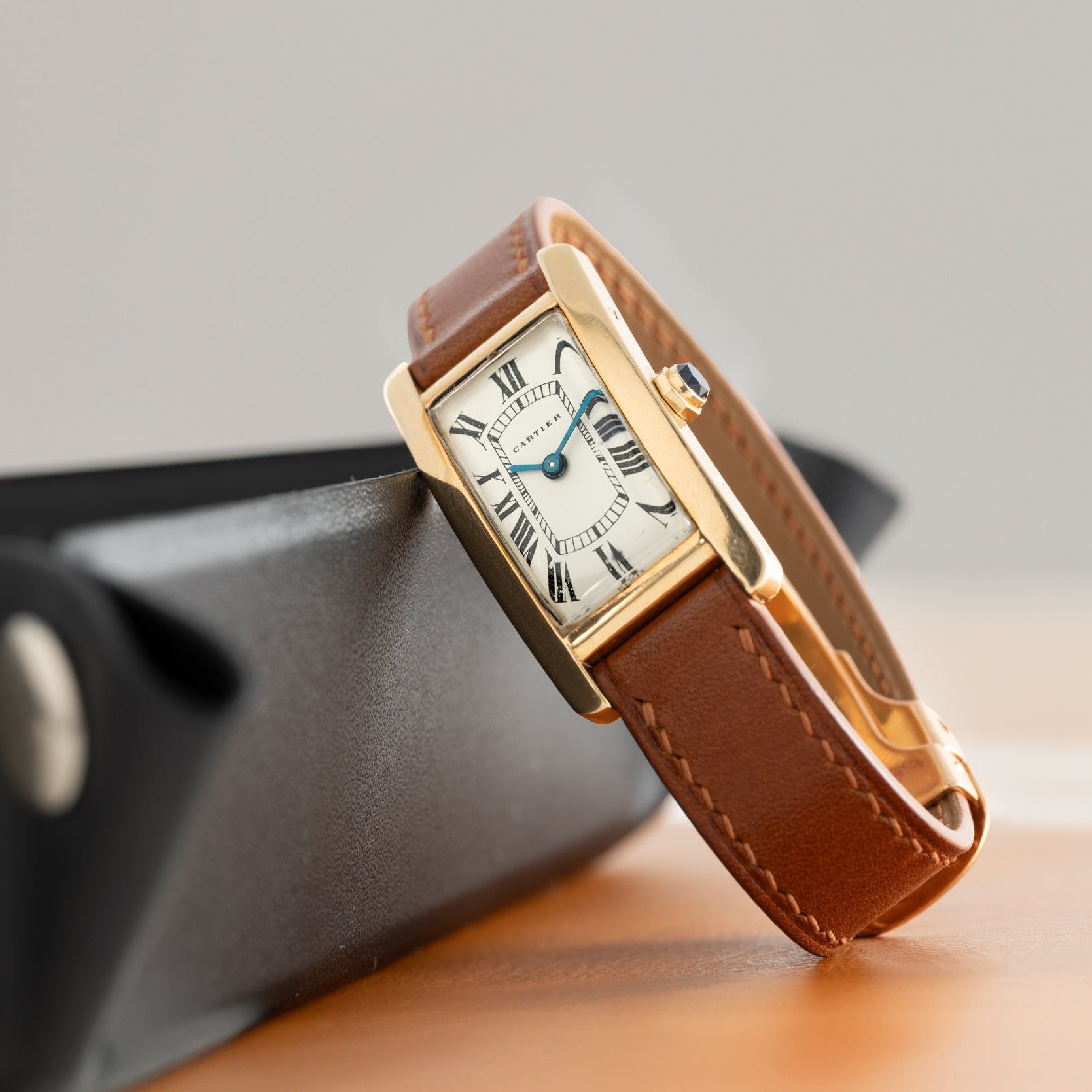 The Cartier Tank Cintrée Ladies watch, featuring a gold case, brown leather band, and Roman numerals, is displayed on a black case.