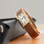 The Cartier Tank Cintrée Ladies watch, featuring a gold case, brown leather band, and Roman numerals, is displayed on a black case.