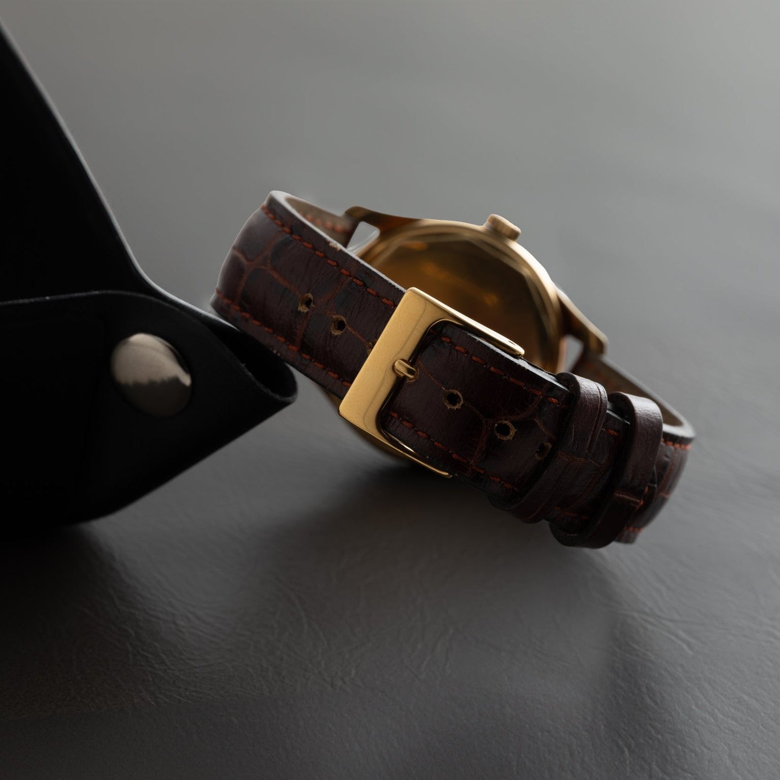 The Patek Philippe Calatrava, a brown leather watch with a gold buckle, rests on a dark surface beside its black case.