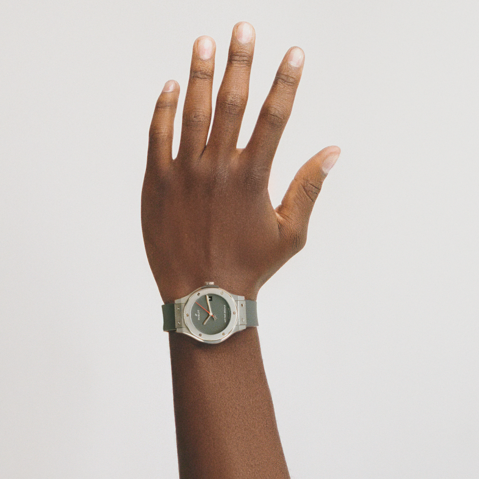 A hand with dark skin is raised against a white background, wearing the Hublot Classic Fusion Titanium Limited Edition for Hodinkee watch, which features a sleek silver strap and a dark face.