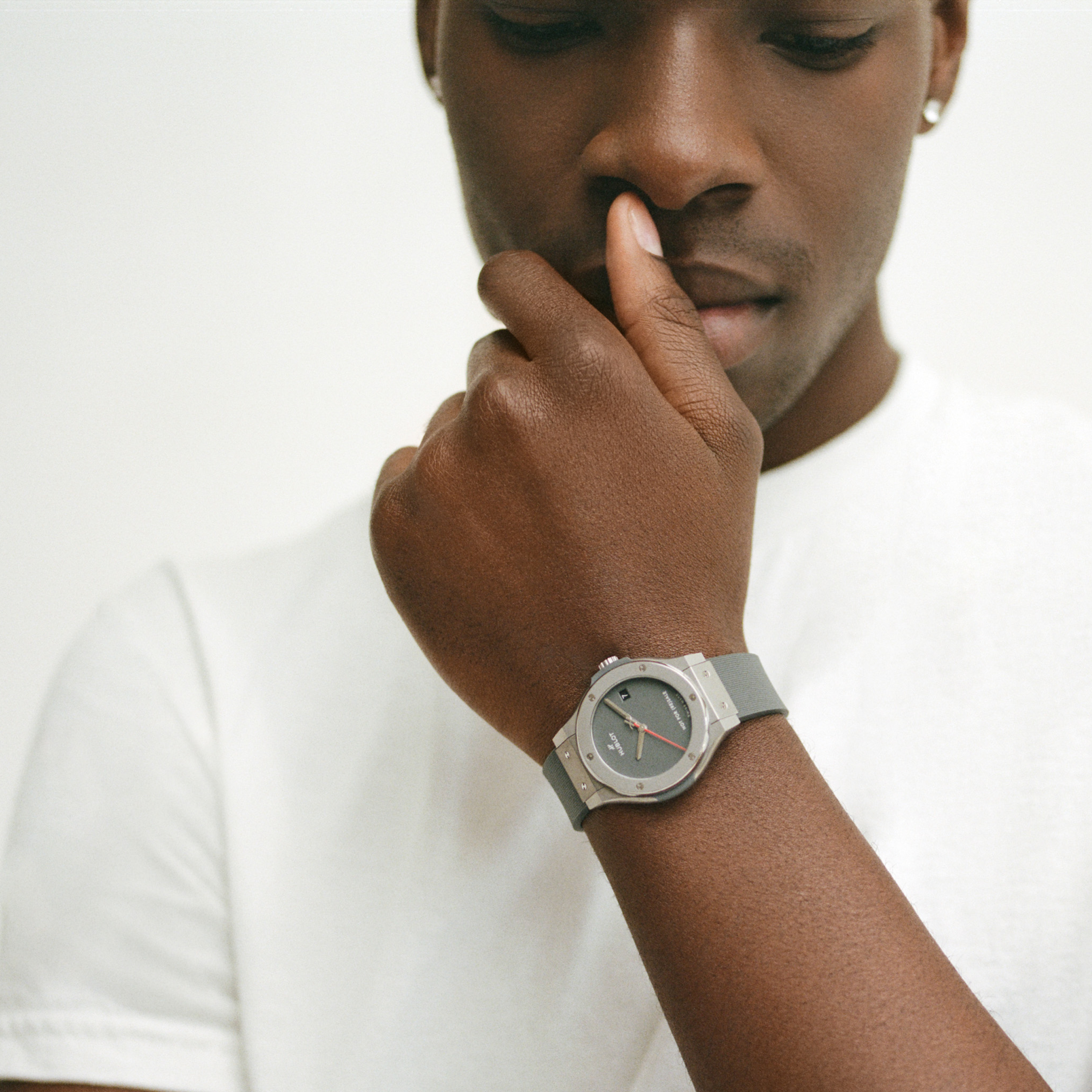 A person in a white t-shirt touches their face, wearing the Hublot Classic Fusion Titanium Limited Edition For Hodinkee wristwatch by Hublot, shown in close-up against a plain background.