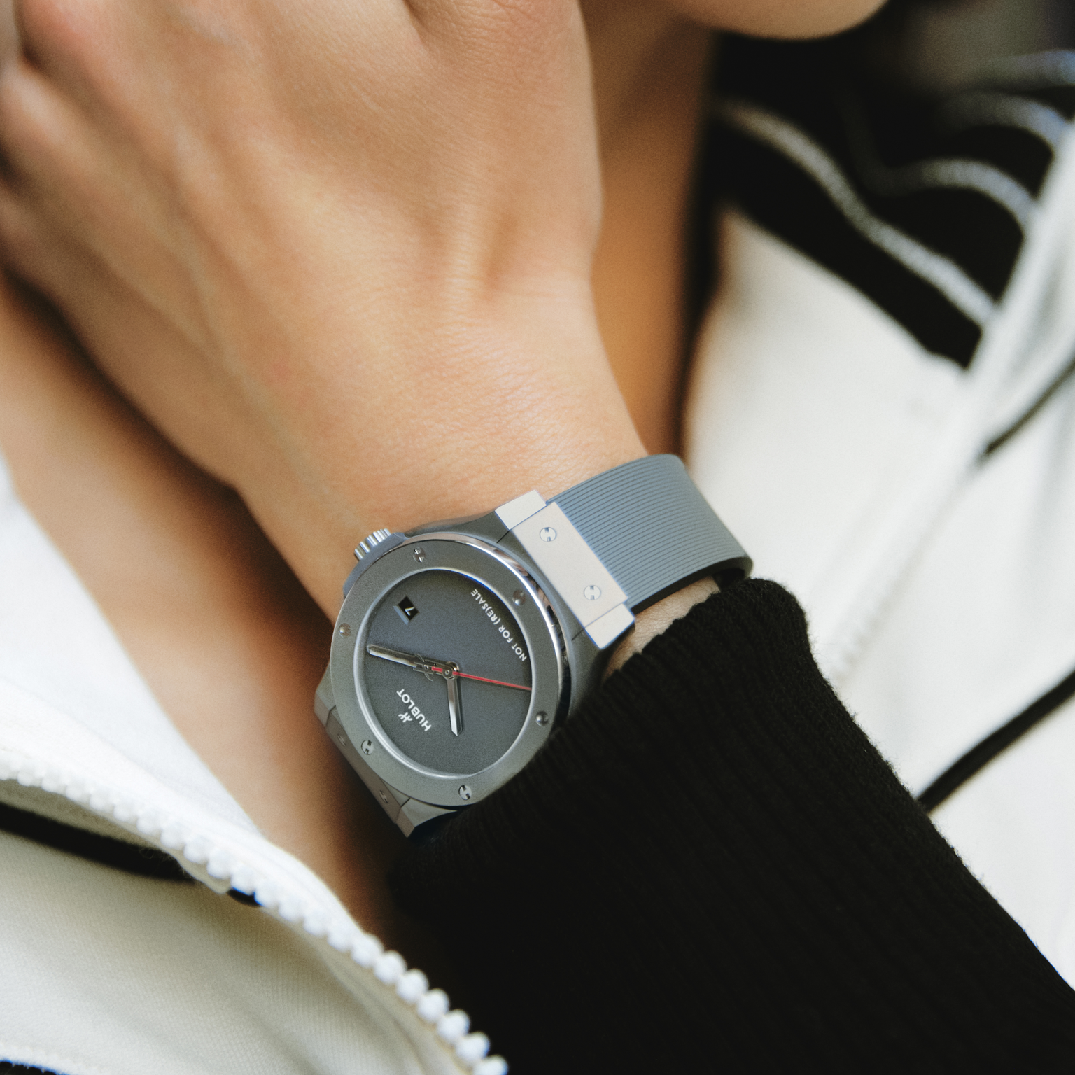A person wearing a Hublot Classic Fusion Titanium Limited Edition For Hodinkee wristwatch with a round face and gray strap, dressed in a white jacket with black trim.