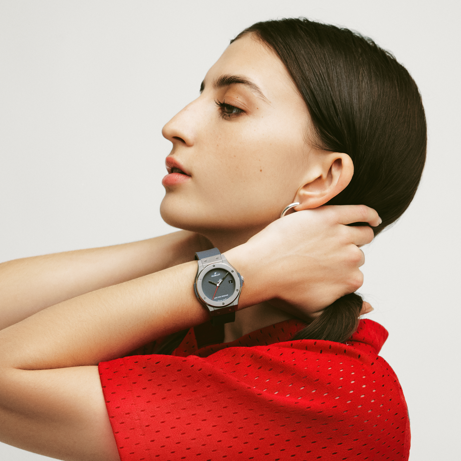 A person in a red shirt adjusts their hair, wearing a Hublot Classic Fusion Titanium Limited Edition For Hodinkee wristwatch and a silver hoop earring, shown in profile against a plain background.