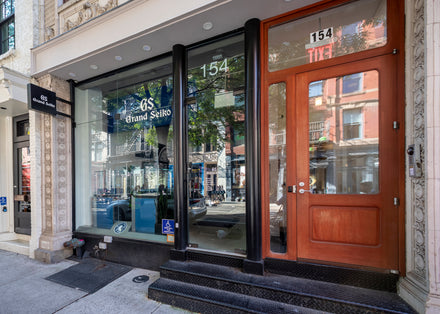 Storefront with large front window displaying Grand Seiko at 154, with a wooden door and street reflection.