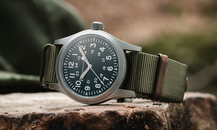 A Hamilton watch with a green khaki fabric strap and a silver tone bezel, sitting outdoors on a rock surface