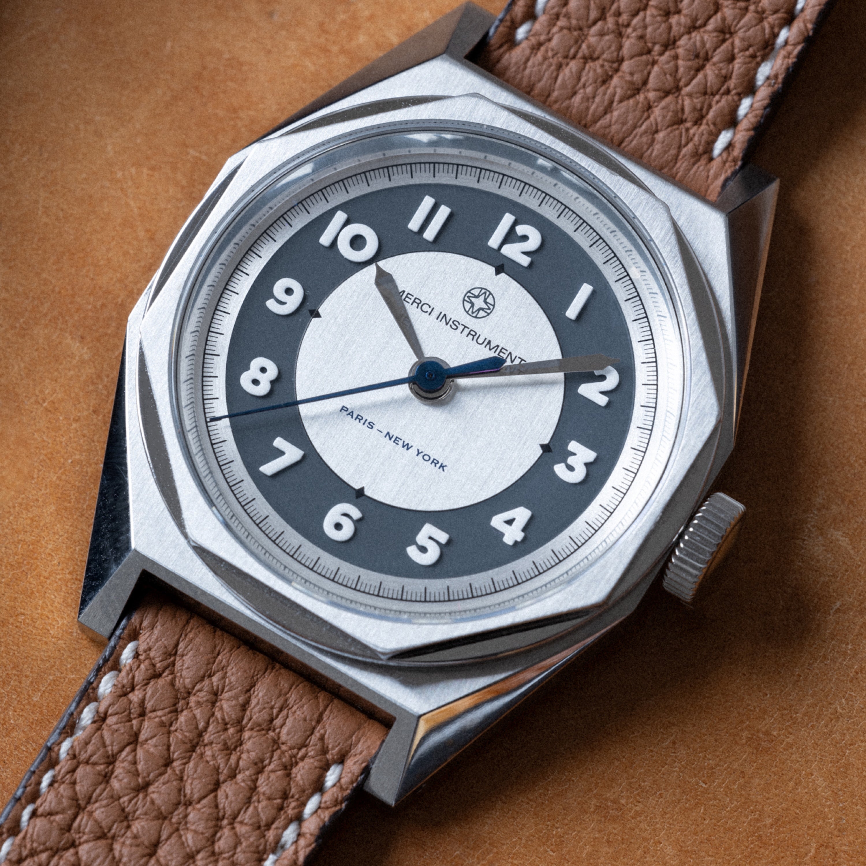 The Merci INSTRUMENTS Beaumarchais H02 Limited Edition For Hodinkee by Merci features a silver case, brown leather strap, and bold white numerals on a gray and white dial.