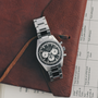The Zenith Chronomaster Original Triple Calendar Limited Edition For Hodinkee, a stainless steel chronograph wristwatch with a black and white dial, rests on a brown leather folder with a printed document partially visible underneath.