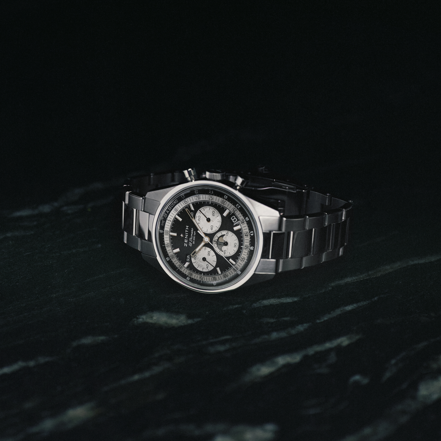 The Zenith Chronomaster Original Triple Calendar Limited Edition for Hodinkee, a stainless steel chronograph with a black dial and three subdials, is showcased against a dark marbled background.