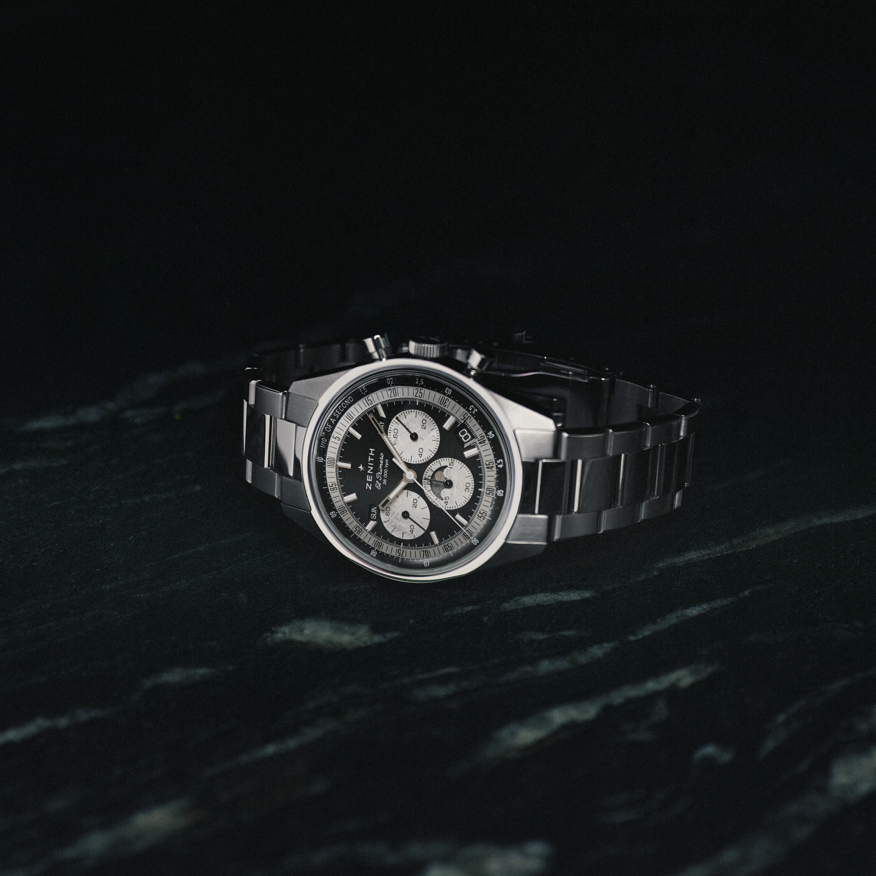 The Zenith Chronomaster Original Triple Calendar Limited Edition for Hodinkee, a stainless steel chronograph with a black dial and three subdials, is showcased against a dark marbled background.