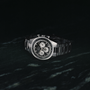 The Zenith Chronomaster Original Triple Calendar Limited Edition for Hodinkee, a stainless steel chronograph with a black dial and three subdials, is showcased against a dark marbled background.