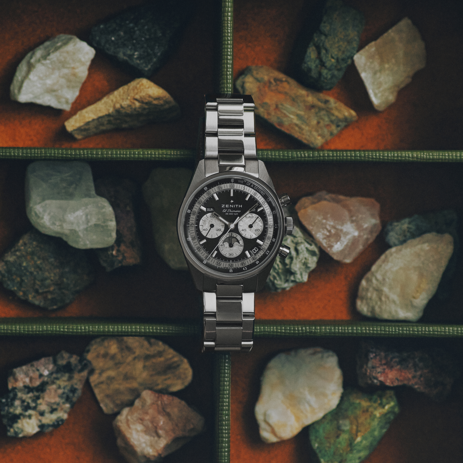 The Zenith Chronomaster Original Triple Calendar Limited Edition For Hodinkee wristwatch is showcased among colorful rocks in a segmented box set against an orange background.