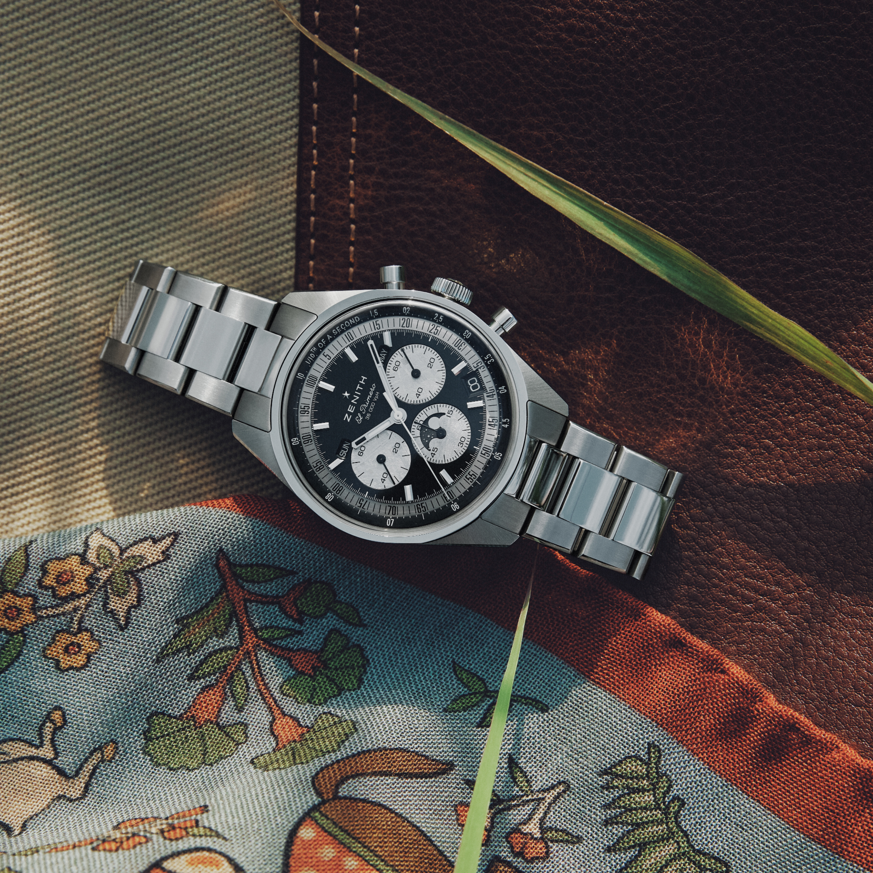 The Zenith Chronomaster Original Triple Calendar Limited Edition For Hodinkee, a stainless steel watch with a black and white dial and modern chronograph subdials, rests on patterned fabric and leather with green grass accents.