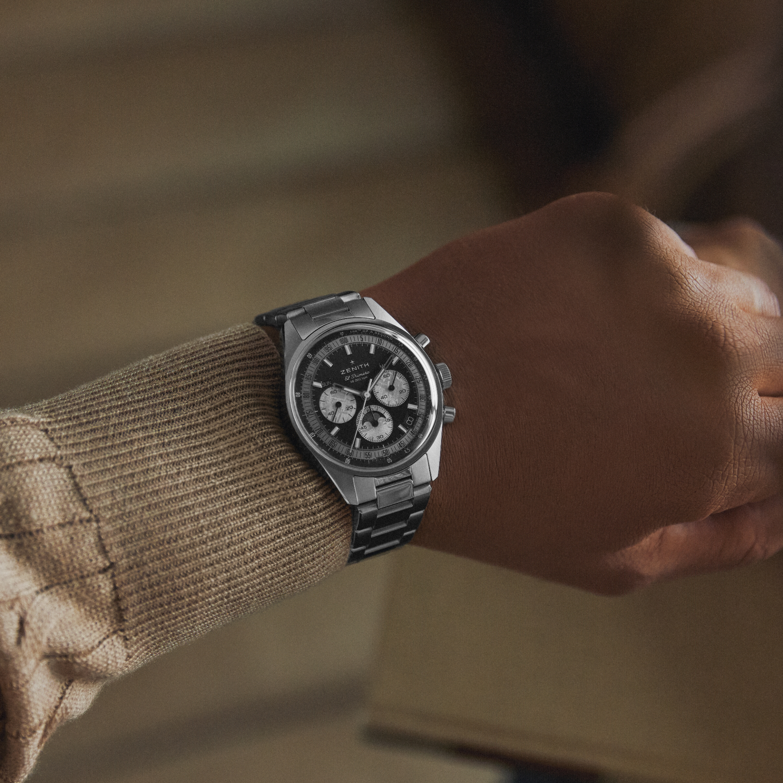 A person wearing a Zenith Chronomaster Original Triple Calendar Limited Edition For Hodinkee, featuring a silver case and black-and-white chronograph dial, dressed in a beige textured sweater and holding a light brown object.