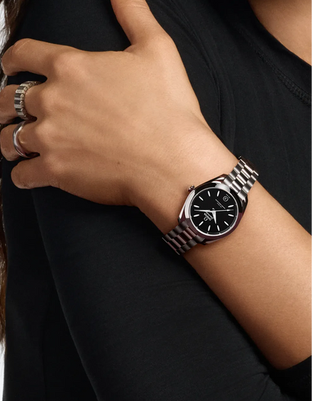 A person wearing a silver wristwatch with a black face and metal band, dressed in a black long-sleeve shirt.