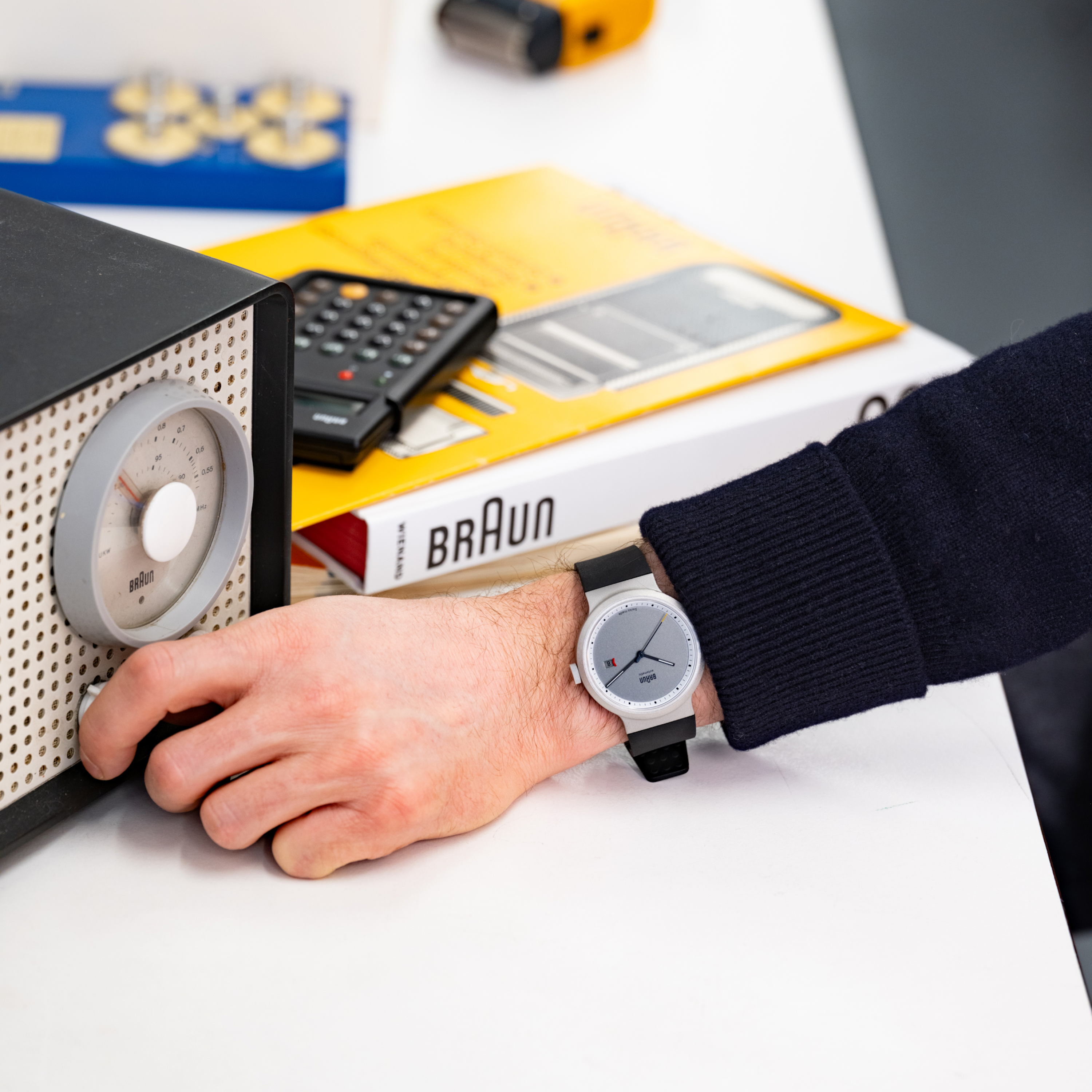 Someone adjusts a vintage Braun radio, wearing the Braun BN0279 Sub-Seconds Limited Edition For Hodinkee watch, with a calculator and book nearby.