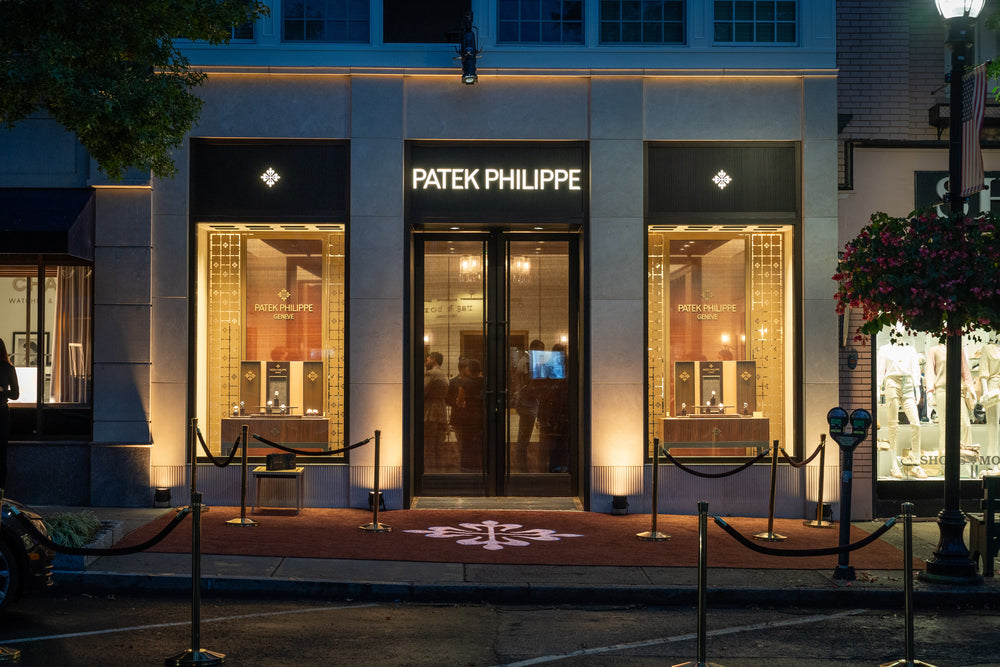 Patek Philippe store exterior at night, warmly lit with displays and a roped entrance.