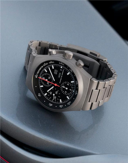 A stainless steel chronograph wristwatch with a black dial and red second hand, resting on a gray surface.