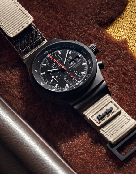 A black chronograph wristwatch with a beige strap resting on a brown textured surface.