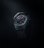 The Chronograph 1 75 Years Porsche Edition by Porsche Design is a black watch with a black strap featuring a red and blue stripe, set against a dark background.